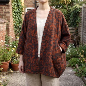 May include: An open-front jacket featuring a dark brown base with a repeating burnt orange floral pattern. The jacket has three-quarter length sleeves, a V-neckline, a button-down front closure, and two side pockets, worn over a white top.