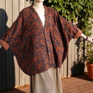 Può includere: Giacca stile kimono con stampa floreale marrone e nera, con scollo a V e chiusura con bottoni. La giacca ha maniche ampie e polsini marroni. La modella indossa un top bianco e una gonna beige. Lo sfondo comprende una recinzione in legno e vegetazione.