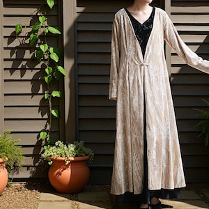 May include: A long, beige velvet coat with a floral pattern and a single clasp closure. The coat is worn over a black top and skirt. The background features a wooden fence, potted plants, and climbing vines.