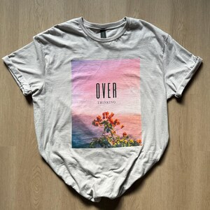 May include: A light gray t-shirt with a graphic print of a sunset with flowers and the text "OVER THINKING".