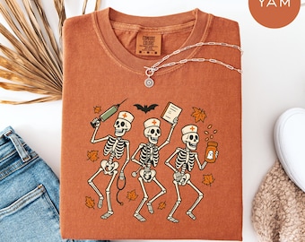以下が含まれることがあります： 錆色のComfort Colors Tシャツ。3人の骸骨看護師が医療品を持っているグラフィックが特徴です。デザインには注射器、医療記録、薬瓶が含まれています。太陽のペンダントが付いたシルバーのネックレスがシャツの上にかけられています。