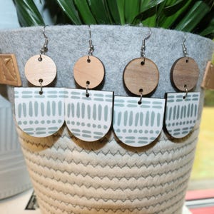 May include: Four pairs of earrings. Each earring features a light wood circle top and a white, semi-circular bottom with a blue-green dotted and dashed pattern. The earrings hang from silver-colored hooks.