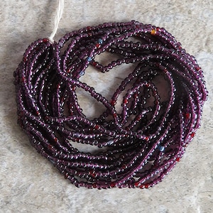 May include: A strand of purple seed beads with a few red and blue beads interspersed. The beads are arranged in a circle and are ready to be used for jewelry making.
