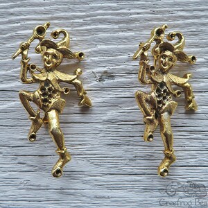 May include: Two gold-toned metal jester figurines with a whimsical design. Each jester is holding a staff and has a bell on their hat.