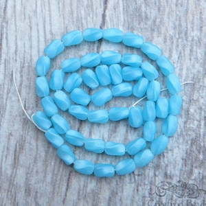 May include: A strand of light blue glass beads with a faceted, slightly irregular shape. The beads are strung on a white thread and are arranged in a circular pattern.