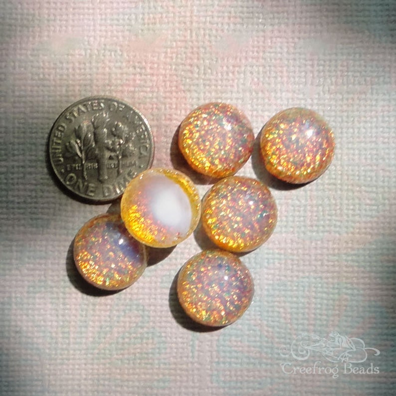 Op de afbeelding: Zeven iriserende glazen cabochons in tinten van goud en wit. De cabochons zijn rond en hebben een glinsterende afwerking.