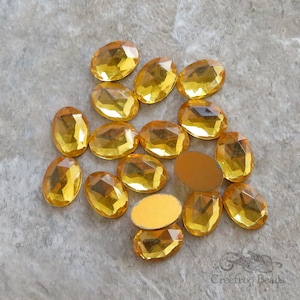 May include: A group of 16 oval-shaped, faceted, clear glass rhinestones with a yellow amber colour. The rhinestones are scattered on a light grey textured surface.