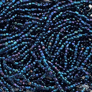 May include: A close-up of a pile of dark blue seed beads with iridescent shimmer. The beads are small and cylindrical, and they are arranged in a random pattern.