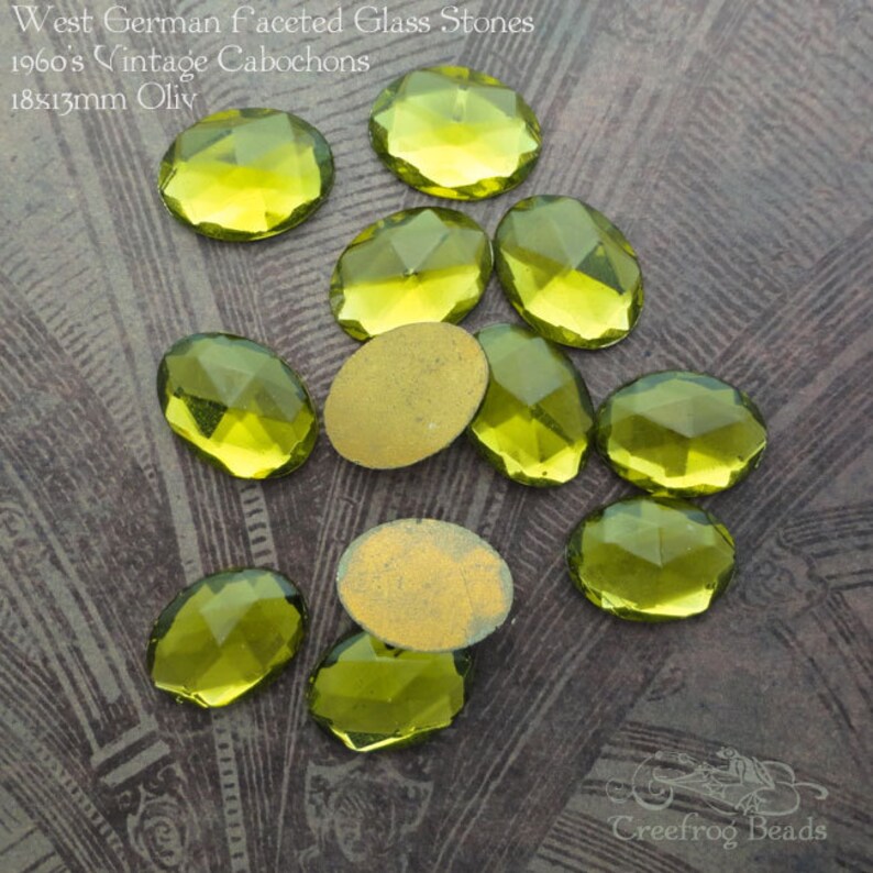 May include: A collection of 18x13mm oval-shaped, faceted glass stones in a light green color. The stones are vintage and from West Germany. The stones are on a textured, gray surface.