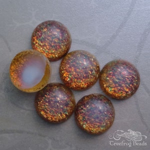 Op de afbeelding: Zes iriserende glazen cabochons in tinten van bruin, oranje en goud met een gespikkeld, getextureerd oppervlak. De cabochons zijn in een halve cirkel op een grijze achtergrond gerangschikt.