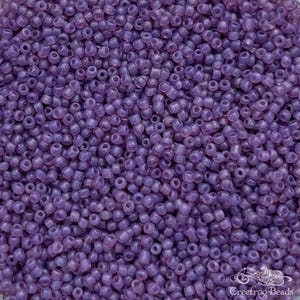 May include: A close-up view of a large quantity of small, opaque, lavender-colored glass beads. The beads are uniform in size and shape, and they have a slightly iridescent sheen.