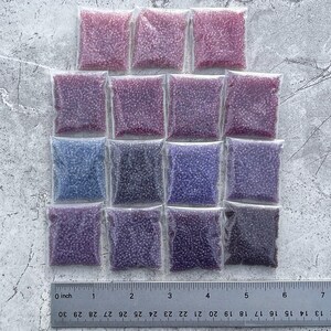 May include: Assortment of 15 small plastic bags filled with seed beads in various shades of purple, pink, and blue. The bags are arranged in three rows of five, with a ruler below them for scale.