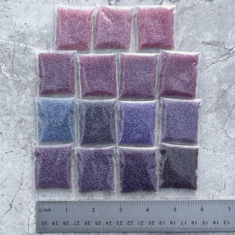 May include: Assortment of 15 small plastic bags filled with seed beads in various shades of purple, pink, and blue. The bags are arranged in three rows of five, with a ruler below them for scale.