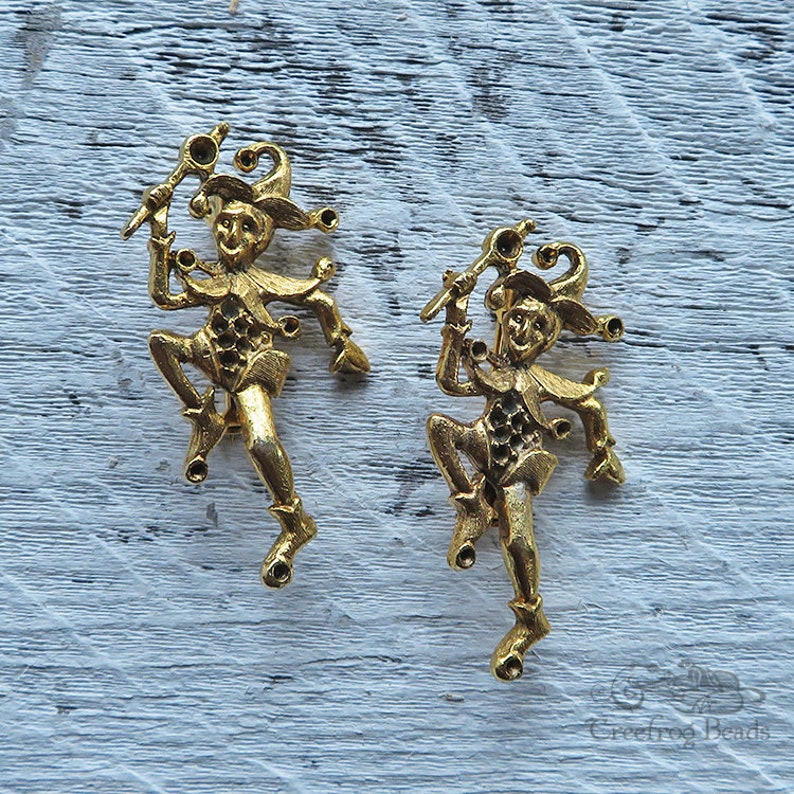 May include: Two gold-toned metal jester figurines with bells and a staff. The jesters are in a dancing pose.
