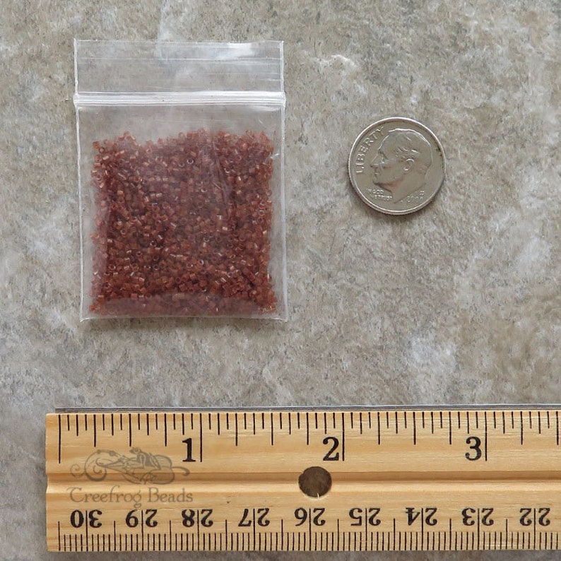 May include: A small clear plastic bag filled with approximately 1 ounce of small, dark red, glass seed beads. The bag is next to a US dime for size comparison. A wooden ruler is in the bottom of the image.