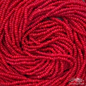 Puede incluir: Primer plano de una hebra de pequeñas cuentas de semillas de color rojo brillante. Las cuentas son redondas y tienen un acabado liso y brillante.