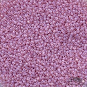 May include: A close-up shot of a pile of small, translucent, light purple seed beads. The beads are cylindrical with rounded edges and a hole running through the center. The beads are scattered, creating a textured surface. The image is well-lit, highlighting the beads' color and shape.