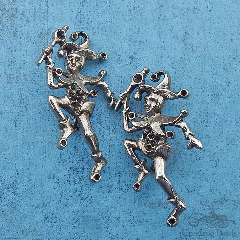 May include: Two silver jester figurines with black accents and a whimsical design. The jesters are holding a staff and are in a dancing pose.