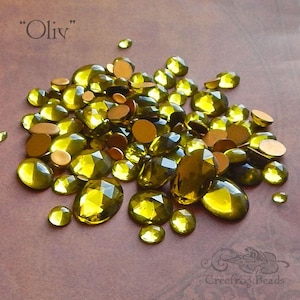 May include: A large pile of olive green glass rhinestones, some with a faceted finish, on a brown surface. "Oliv" is written in white text at the top of the image.