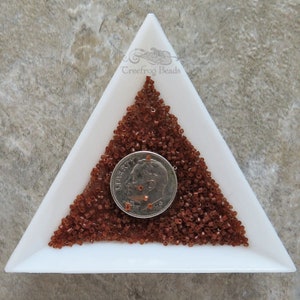 May include: A white triangular tray filled with a pile of small, brown glass seed beads. A US dime coin is placed on top of the beads for size comparison.