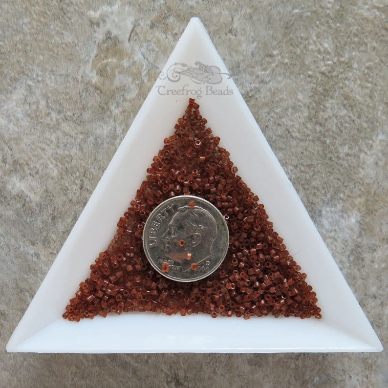 May include: A white triangular tray filled with a pile of small, brown glass seed beads. A US dime coin is placed on top of the beads for size comparison.