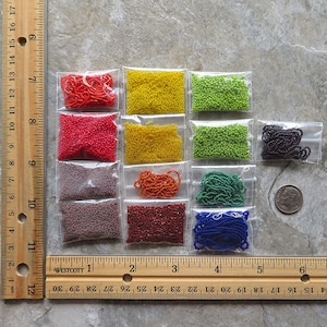 May include: Assortment of small glass beads in various colors, including red, orange, yellow, green, blue, black, and brown. The beads are packaged in small clear plastic bags and are arranged in rows on a wooden ruler.