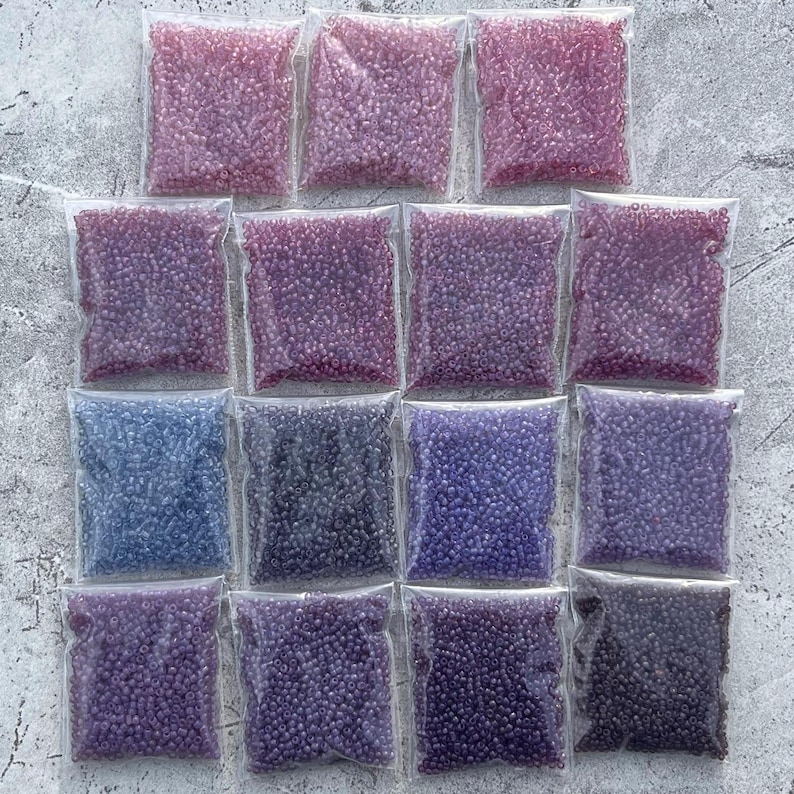 May include: Assortment of 15 small bags of seed beads in various shades of purple and blue. The beads are packaged in clear plastic bags.