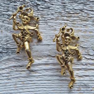 May include: Two gold-toned jester figurines with a whimsical design. Each jester is holding a staff with a ball on top and has a jester hat with bells. The jesters are standing on a wooden surface.
