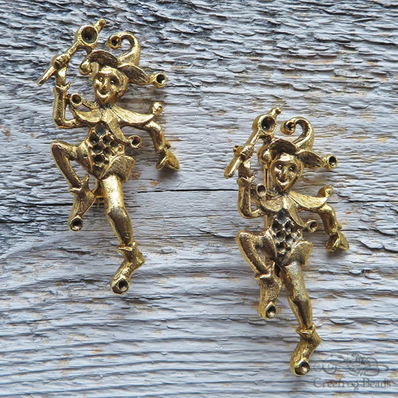 May include: Two gold-toned jester figurines with a whimsical design. Each jester is holding a staff with a ball on top and has a jester hat with bells. The jesters are standing on a wooden surface.