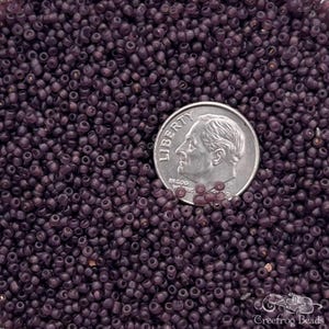 May include: A close-up view of a large quantity of small, round, opaque purple glass beads. A US dime is included for scale.