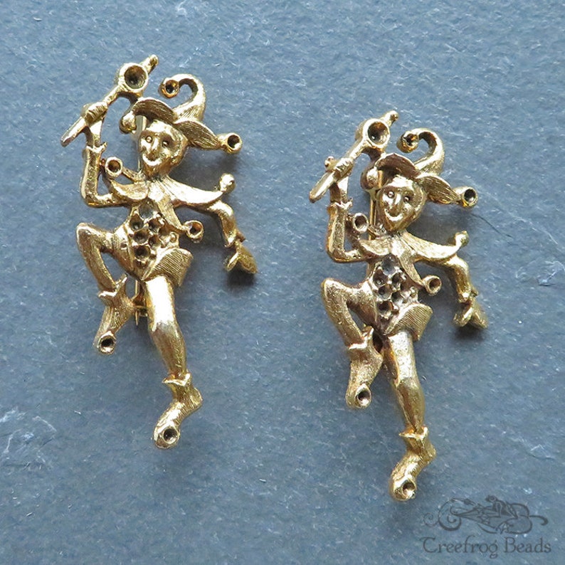 May include: Two gold-tone metal jester figurines with a whimsical design. Each jester is holding a staff and has a jester hat with bells.