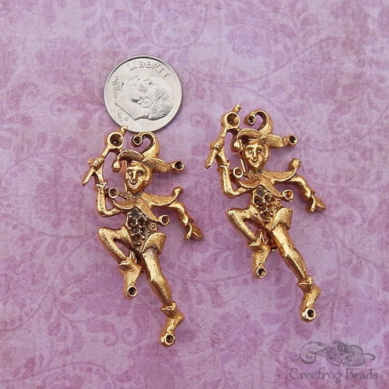 May include: Two gold-tone jester figurines with rhinestones. Each jester is holding a staff and has a whimsical expression.