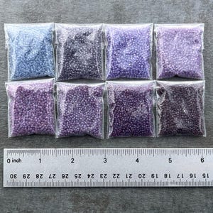 May include: Eight small bags of seed beads, arranged in two rows of four. The beads are in shades of purple and blue, with a ruler for scale.