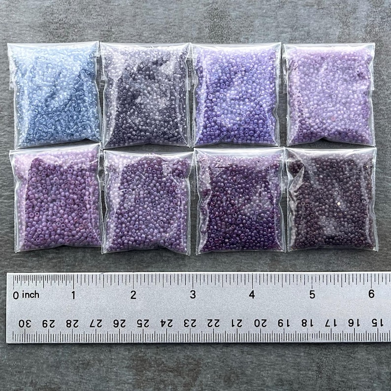 May include: Eight small bags of seed beads, arranged in two rows of four. The beads are in shades of purple and blue, with a ruler for scale.