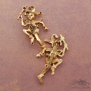 May include: Gold-toned metal brooch featuring two jester figures with rhinestones. The jester figures are in a dancing pose. The brooch has a pin back.