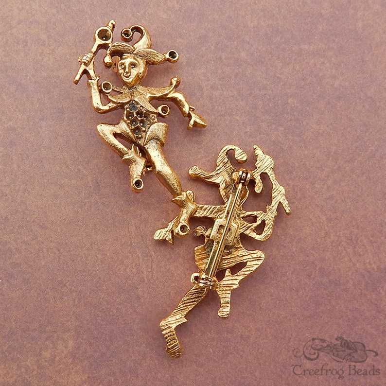 May include: Gold-toned metal brooch featuring two jester figures with rhinestones. The jester figures are in a dancing pose. The brooch has a pin back.