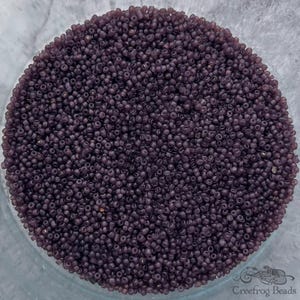 May include: A close-up view of a bowl filled with small, dark purple glass beads. The beads are uniform in size and shape, creating a textured surface.