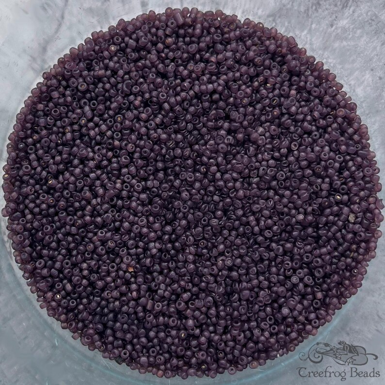 May include: A close-up view of a bowl filled with small, dark purple glass beads. The beads are uniform in size and shape, creating a textured surface.