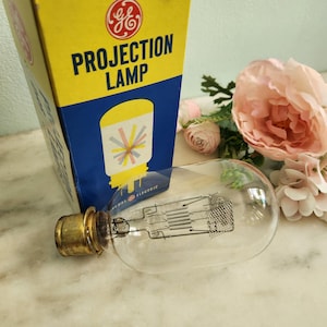 May include: A vintage General Electric projection lamp with a clear glass bulb and a brass base. The lamp is next to its original box, which is yellow and blue with the words "PROJECTION LAMP" and a graphic of a lamp. Pink flowers are in the background.