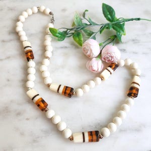 May include: A necklace featuring a series of round, matte off-white beads interspersed with cylindrical beads that combine off-white and translucent amber-colored segments with dark brown stripes. Small metallic gold-toned spacer beads are present.