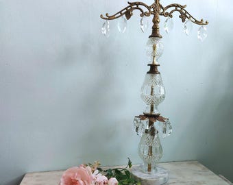 May include: An ornate, vintage-style lamp with a brass finish and crystal accents. The lamp features a decorative top with hanging crystals, a cut-glass body, and a marble base. A pink rose and smaller pink flowers are on the table.