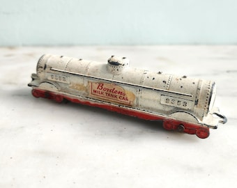 May include: Vintage toy milk tank car, cream-colored with red accents. The words "Borden's Milk Tank Car" are visible on the side. The toy has the number "9353" printed on it. The wheels and undercarriage are red.