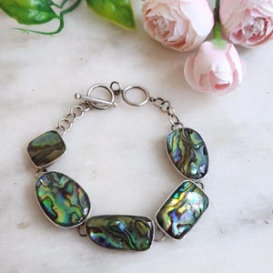 May include: A bracelet featuring five polished abalone shell segments with iridescent green, blue, and purple hues, connected by silver-toned metal links and secured with a circular toggle clasp. The shell pieces vary in rectangular and oval shapes.