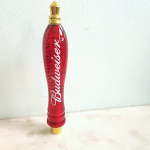 May include: A red Budweiser tap handle with a gold-colored top and base. The handle is a deep red with the Budweiser logo in white script. The handle has a textured, ribbed design.