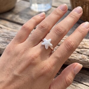 May include: A silver-toned ring featuring a textured, five-pointed starfish design. The starfish has a bumpy, realistic surface detail and is centred on a smooth, polished metal band that encircles the finger, creating a nautical-inspired accessory.