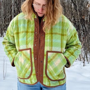 May include: A green and white plaid quilted jacket with brown trim and pockets. The jacket has a button-up front and is worn over a dark shirt and blue jeans. The background is a snowy landscape with bare trees.