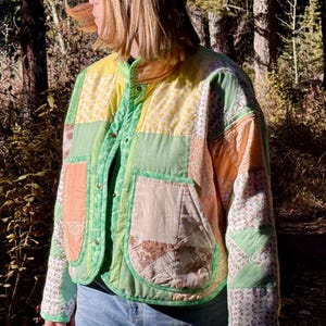 May include: A colourful patchwork jacket in shades of green, yellow, and orange. The jacket features a button-up front and two pockets. The sleeves have a floral pattern. The jacket is worn in a forest setting.