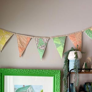 May include: A framed watercolour painting of a house with a green frame hangs on a grey wall, below a garland of colourful fabric pennants. A black metal shelf displays various decorative items and plants.