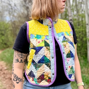 May include: A quilted waistcoat featuring a patchwork design of various triangular fabric patterns in shades of yellow, green, blue, and cream. The waistcoat has a high collar, a purple fabric trim along the edges, and a front closure with metal snap buttons.