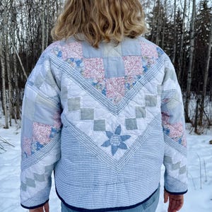 May include: A quilted jacket with a patchwork design. The jacket features a mix of light blue, pink floral, and gray square patterns. The jacket has a blue gingham pattern on the lower back and a blue flower design. The jacket is worn over blue jeans.